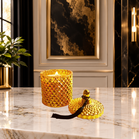 A textured gold glass candle jar, the LéShay Candles Blessed Home Luxury Diamond Jar Candle with a lit wood wick flame sits on a polished marble surface next to its matching gold lid, which has a black tassel. The background features elegant interior decor, including a gold-framed artwork and a black curtain.